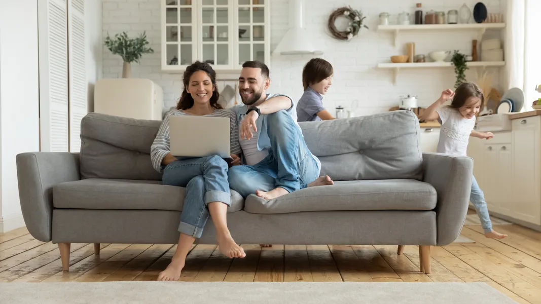 A husband and wife, smiling,sitting on a couch in their home looking at a computer while kids run in the background.